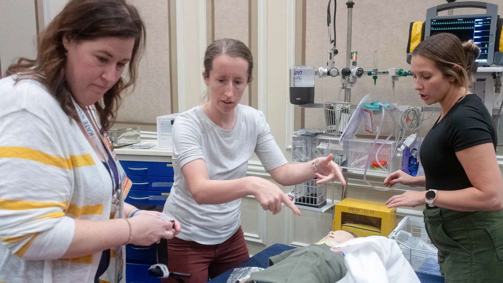 Three healthcare professionals participate in a hands-on training session using a pediatric medical manikin, surrounded by medical equipment in a simulation room.