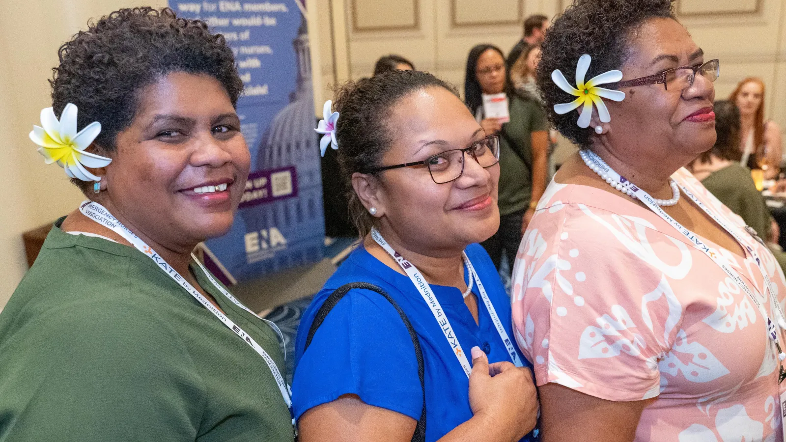 Three women wearing floral hair accessories and ENA lanyards smile and pose together at a conference meet-and-greet event, with attendees and a promotional banner in the background.