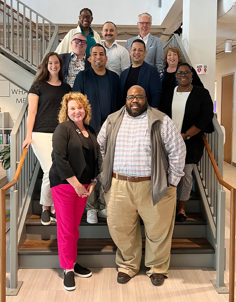 Group photo of a diverse team posing together on a staircase in a modern office space. The individuals are dressed in business casual attire, smiling and looking at the camera.