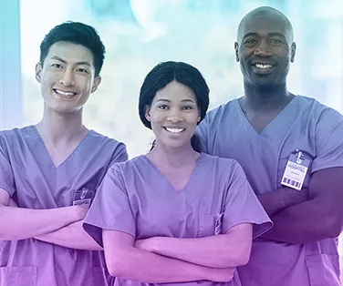Group of three diverse healthcare professionals in scrubs, smiling with arms crossed, standing in a bright medical setting.