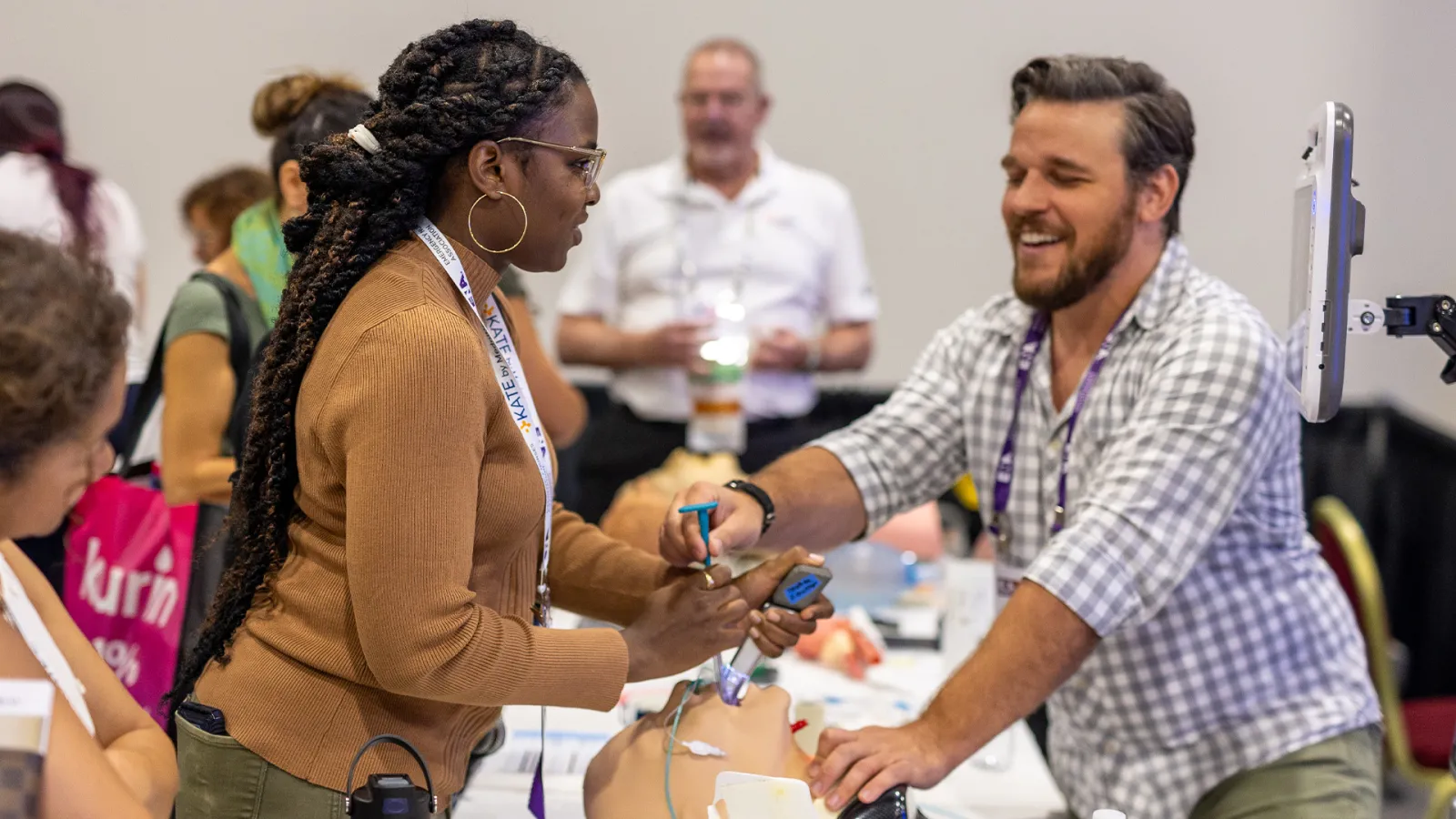 A woman practices intubation on a medical manikin while receiving guidance from a smiling instructor at a hands-on skills station during a health care conference.