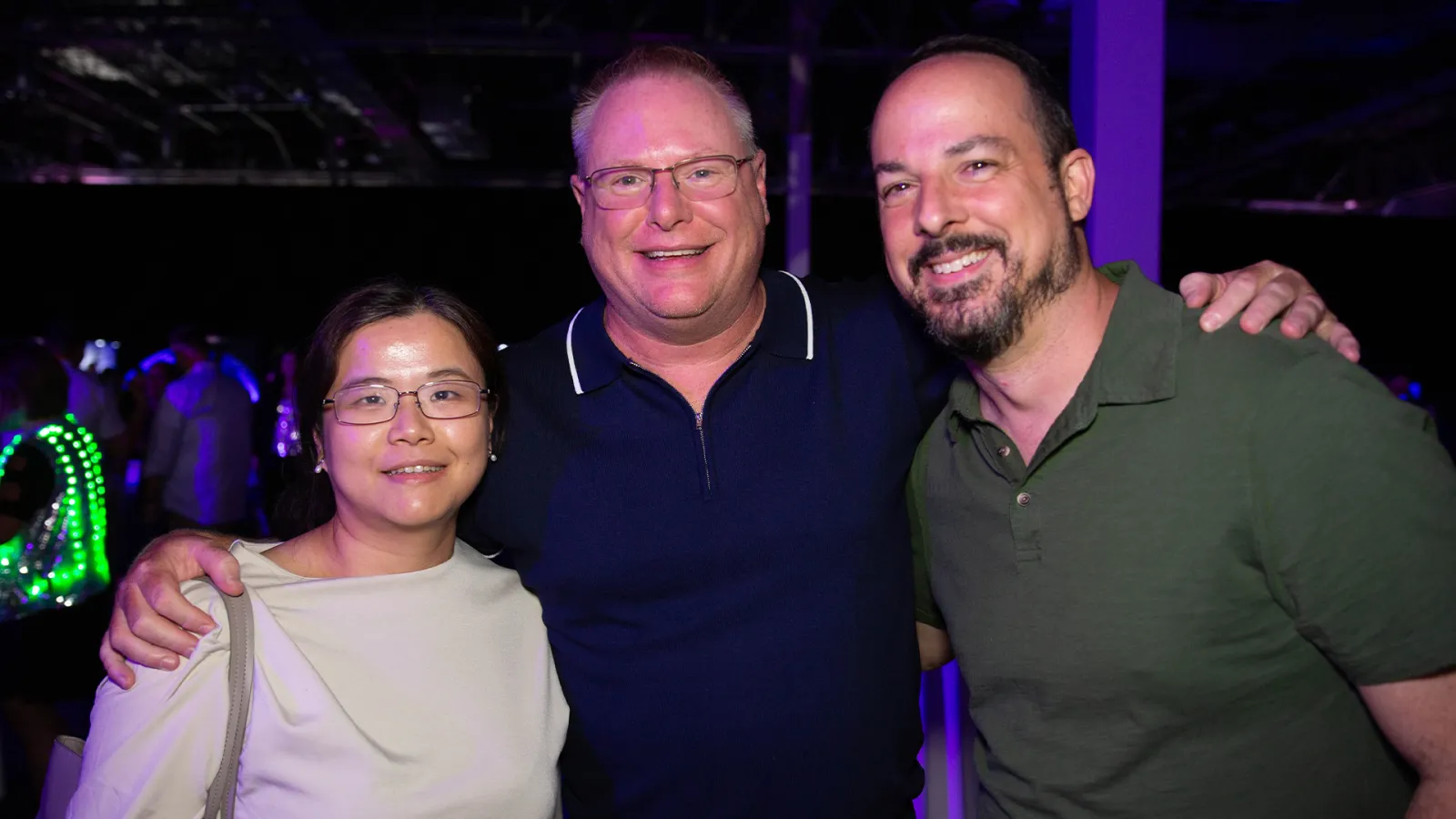 Three attendees smile and pose together under colorful lighting at a lively welcome party, with glowing accessories and other guests in the background.