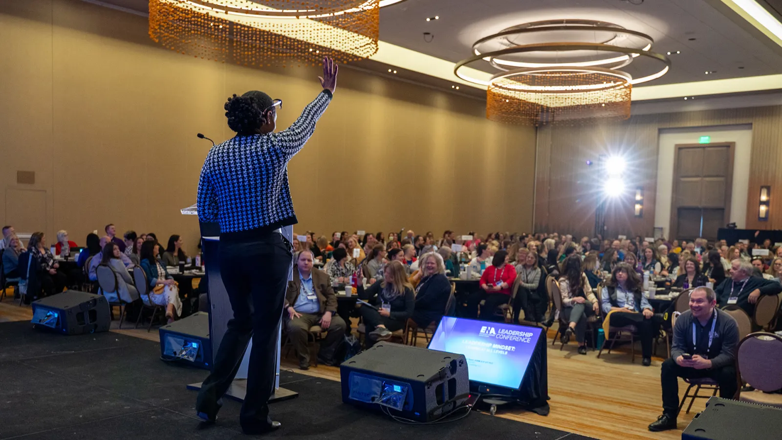 A speaker stands on stage addressing a large audience at the ENA Leadership Conference. The presenter, wearing a black and white patterned jacket, gestures toward the crowd as attendees seated at round tables listen attentively under modern circular light fixtures.