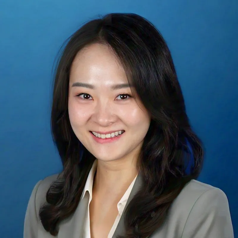 Headshot of a woman with long dark hair, wearing a light gray blazer over a white blouse, smiling warmly against a blue background.