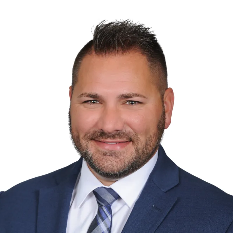 Professional headshot of a man in a navy suit and plaid tie, smiling in front of a white background.