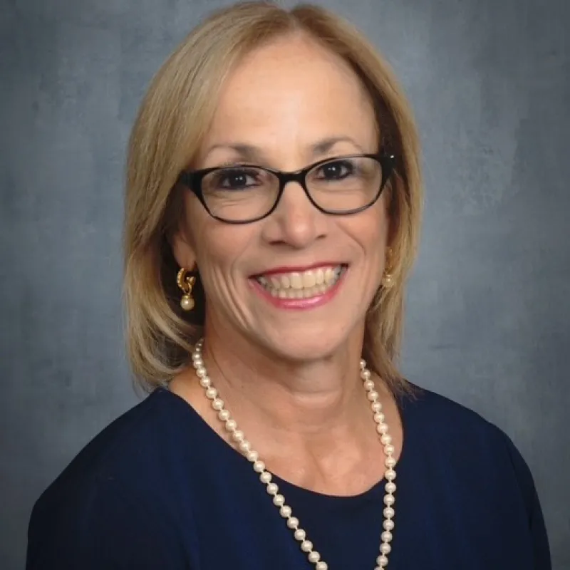 Professional headshot of a woman wearing glasses, a navy blouse, and a pearl necklace, smiling in front of a gray background.