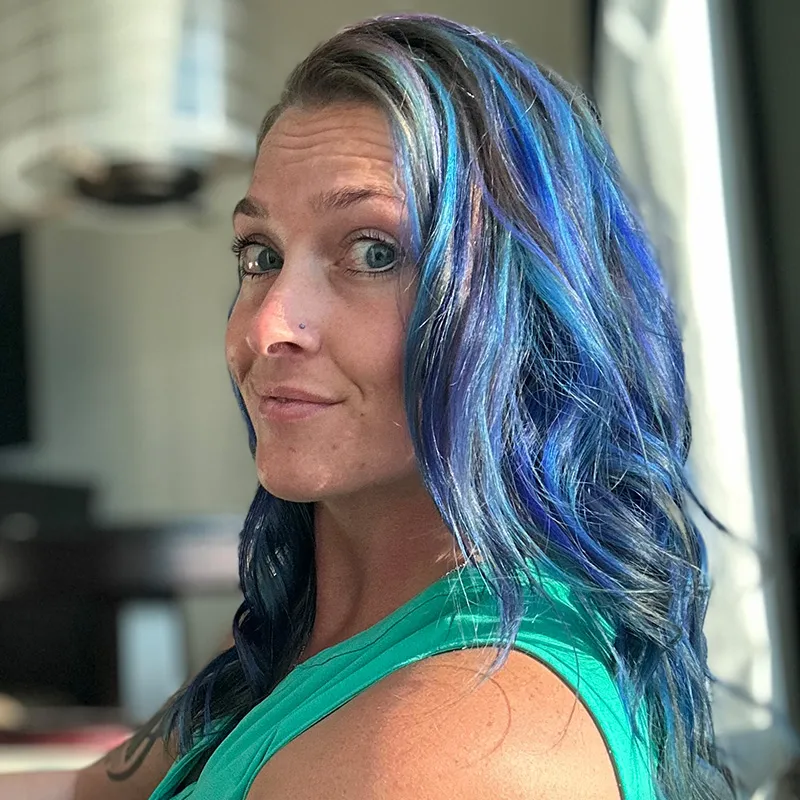 Woman with long, wavy blue and purple hair looking toward the camera with a slight smile, wearing a green sleeveless top in a well-lit indoor setting.