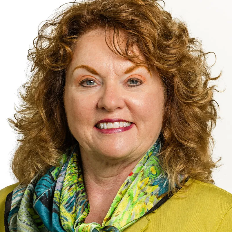 Professional headshot of a woman with curly auburn hair, wearing a green blazer and a colorful patterned scarf, smiling against a white background.