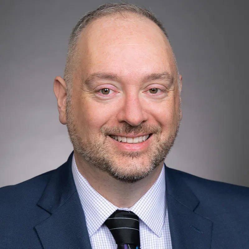 Professional headshot of a smiling man with short hair and a trimmed beard, wearing a navy suit, checkered shirt, and striped tie, against a neutral gray background.