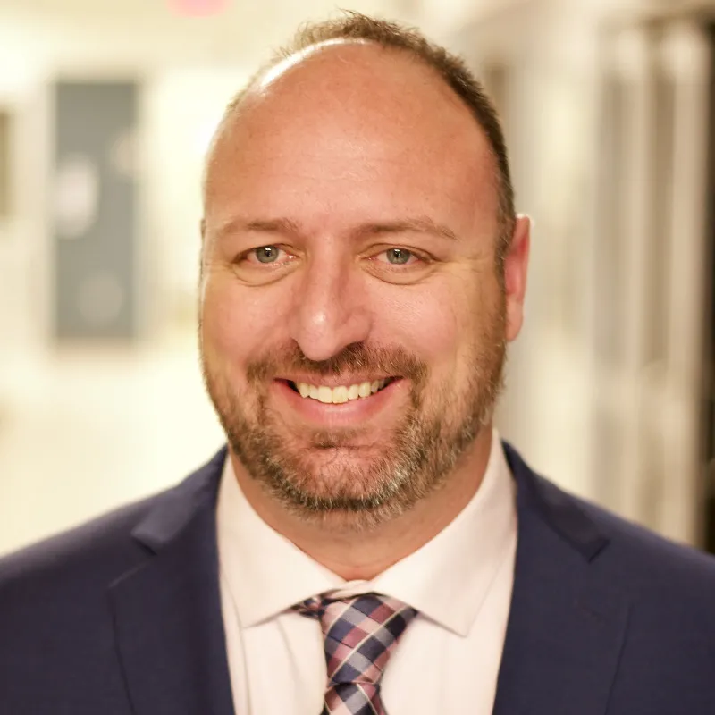 "Professional headshot of a man with a beard and short hair, wearing a navy blue suit, white shirt, and plaid tie, smiling in a brightly lit hallway.
