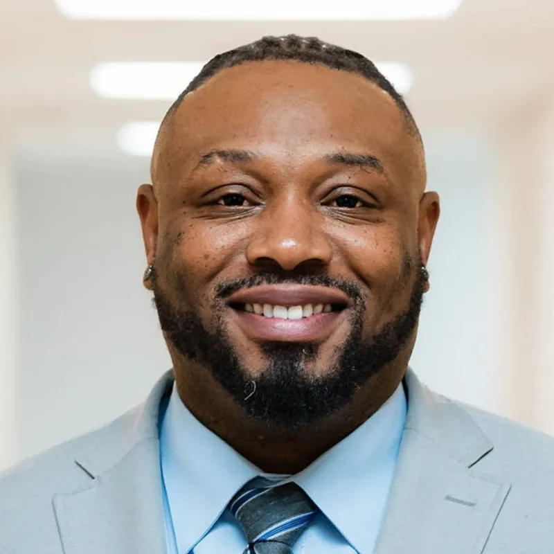 Rahkeem Francis, smiling in a professional headshot, wearing a light gray suit jacket, light blue dress shirt, and a striped blue tie, standing in a brightly lit indoor hallway.