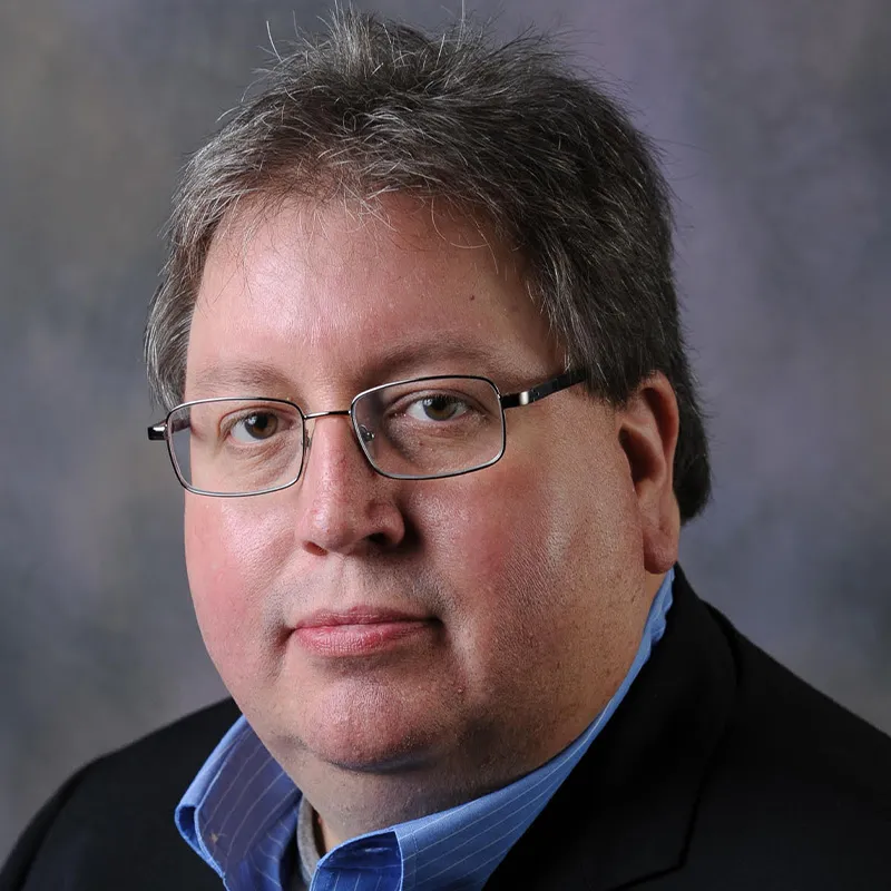 Professional headshot of a man wearing glasses, a dark blazer, and a blue collared shirt, posed against a neutral gray background.