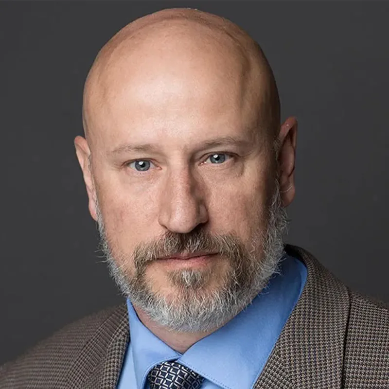 Professional headshot of a man with a bald head and gray beard wearing a brown suit jacket, blue dress shirt, and patterned tie against a dark gray background.