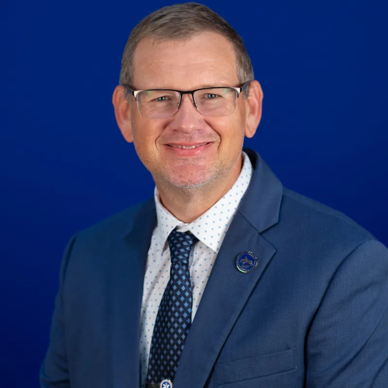 Professional headshot of a man wearing glasses, a blue suit jacket, white patterned shirt, and blue tie, smiling in front of a solid blue background.