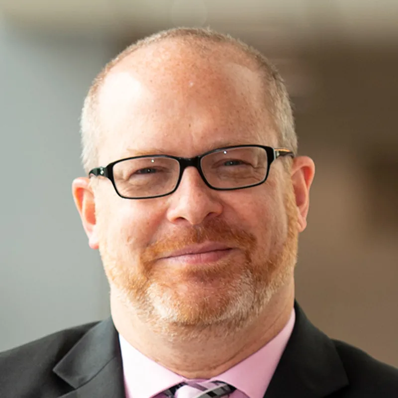 Professional headshot of a man wearing glasses, a black suit, pink shirt, and patterned tie, smiling slightly against a softly blurred indoor background.