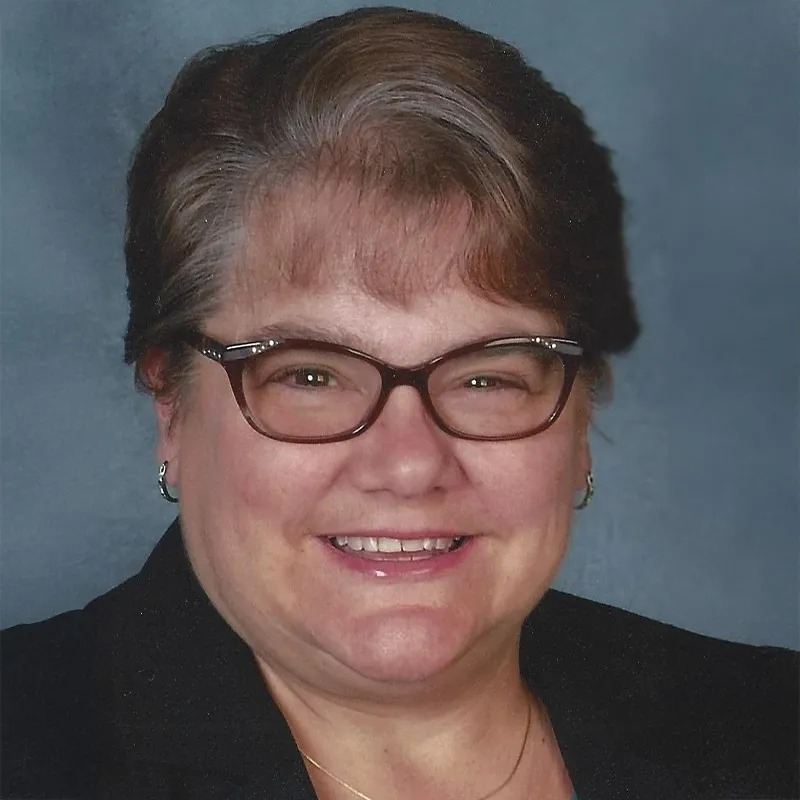 Professional headshot of a smiling woman wearing glasses, a dark blazer, and a patterned blouse, set against a gray-blue background.