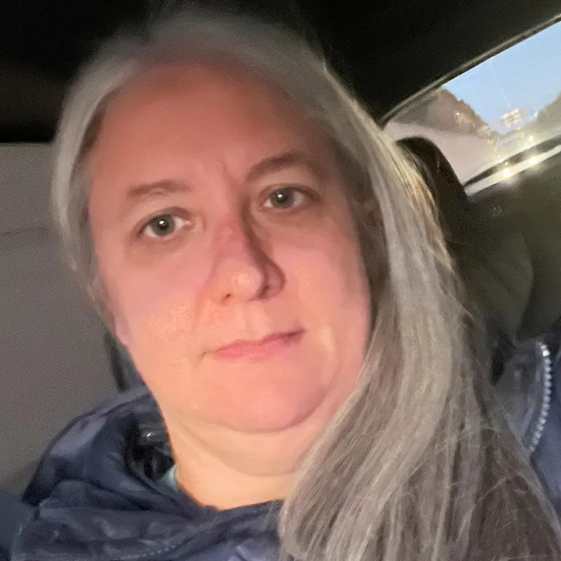 Casual portrait of a woman with long gray hair wearing a dark jacket, sitting inside a car with daylight visible through the rear window.