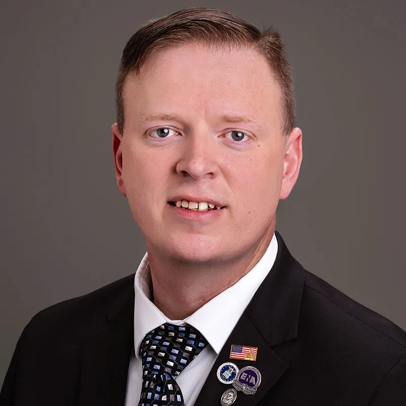 Professional headshot of a man with short light brown hair wearing a black suit jacket, white dress shirt, and patterned tie, facing the camera against a neutral gray background, with an American flag pin and ENA lapel pins on his jacket.