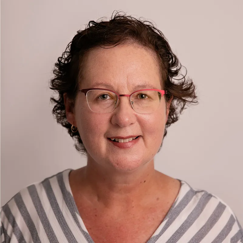 Headshot of a woman with short, curly brown hair wearing red-framed glasses and a striped top, smiling softly against a light neutral background.