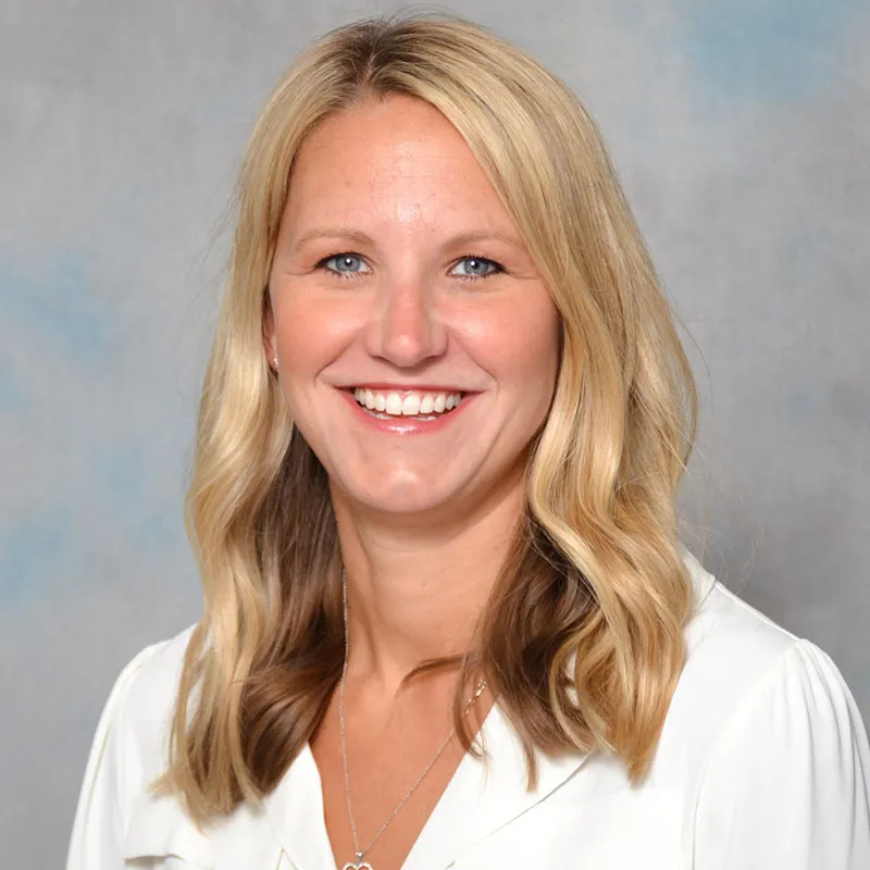 Professional headshot of a smiling woman with shoulder-length blonde hair, wearing a white blouse and a necklace, facing the camera against a soft neutral studio background.