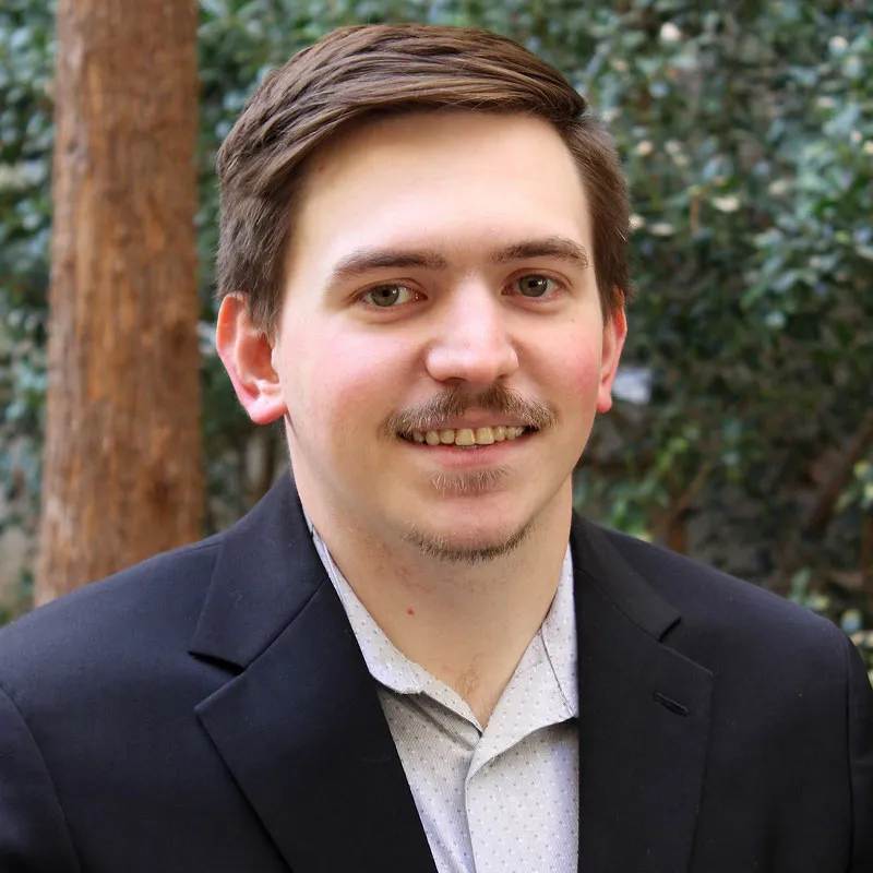 Headshot of a man with short brown hair and a trimmed mustache, wearing a dark blazer over a light patterned shirt, smiling slightly outdoors with greenery and a tree trunk in the background.