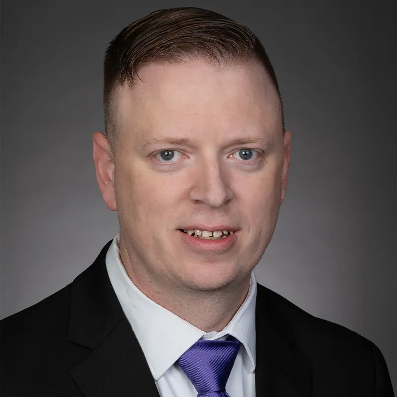 Professional headshot of a man wearing a dark suit jacket, white shirt, and purple tie, with neatly styled hair, facing the camera against a neutral gray background.