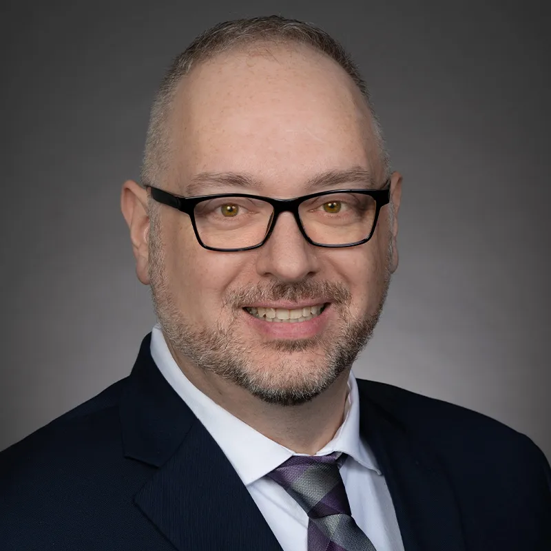 Professional headshot of a smiling person wearing black rectangular glasses, a dark suit jacket, white dress shirt, and patterned tie, photographed against a neutral gray background.