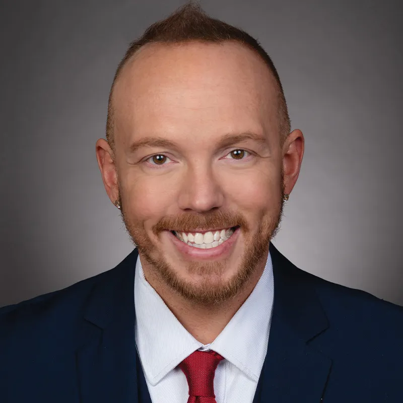 Professional headshot of a smiling person with a closely shaved hairstyle and trimmed beard, wearing a dark suit jacket, white shirt, and red tie against a neutral gray background.