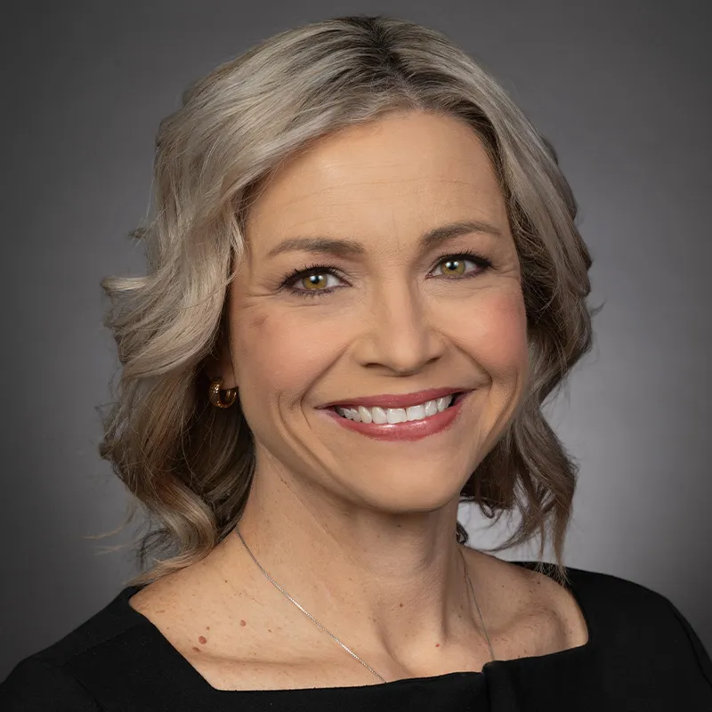 Professional headshot of a woman with shoulder-length gray-blonde hair, wearing a black top and small earrings, smiling against a neutral gray background.