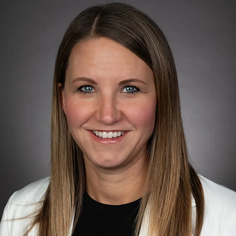 Professional headshot of a woman with long, straight light brown hair, wearing a white jacket over a dark top, smiling against a neutral gray background.