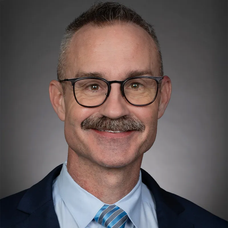 Professional headshot of a middle-aged man wearing glasses, a suit jacket, light blue shirt, and striped tie, smiling against a neutral gray background.