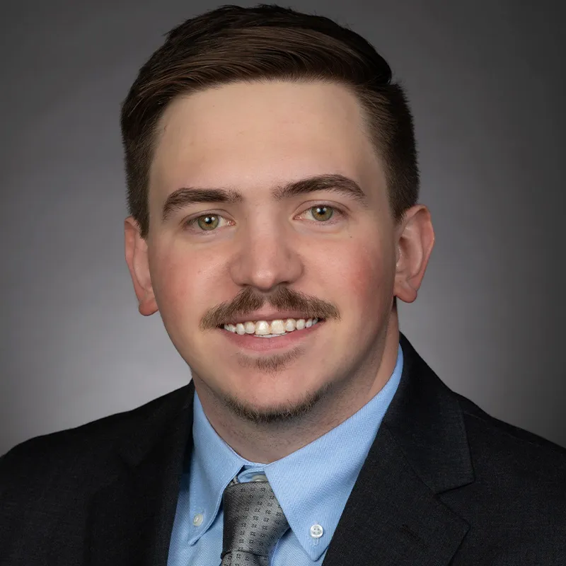 Professional headshot of a man with short brown hair and a trimmed mustache, wearing a dark suit jacket, light blue shirt, and gray tie, smiling against a neutral gray background.