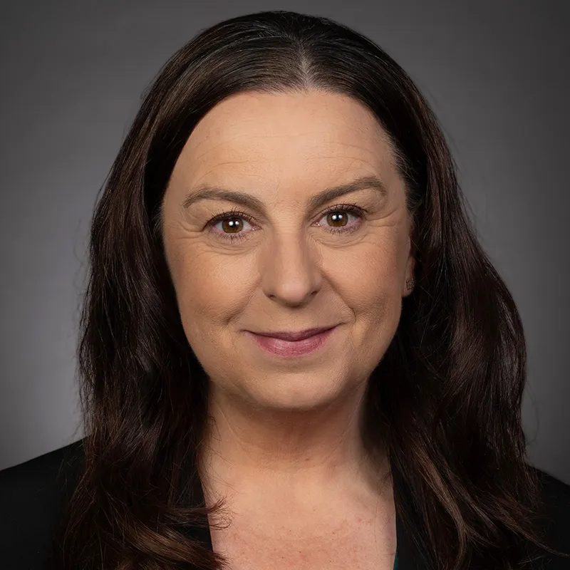 Professional headshot of a person with long dark hair and a subtle smile, wearing a dark blazer, photographed against a neutral gray background.