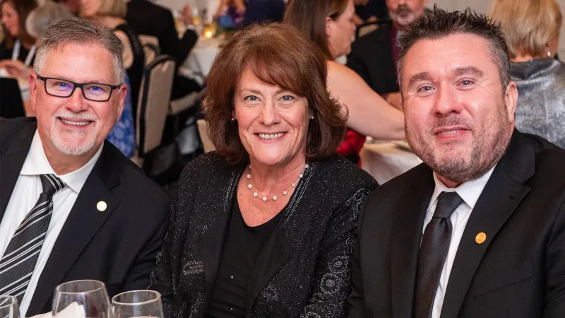 Three professionally dressed attendees smile for a photo during a formal dinner event, likely an awards or recognition ceremony, with other guests seated at tables in the background.