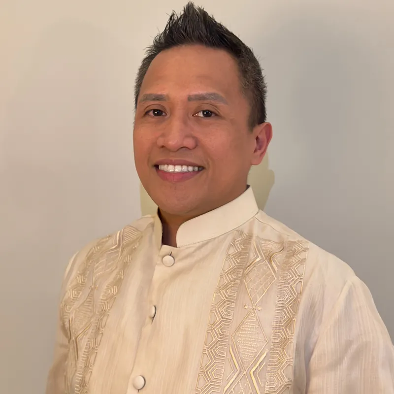 Professional headshot of a man smiling and wearing a traditional embroidered cream-colored barong tagalog, posed against a neutral background.
