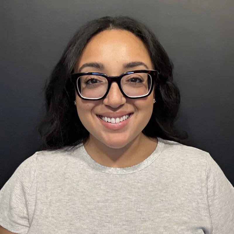 Portrait of a smiling woman wearing glasses and a light gray top, standing in front of a dark background.