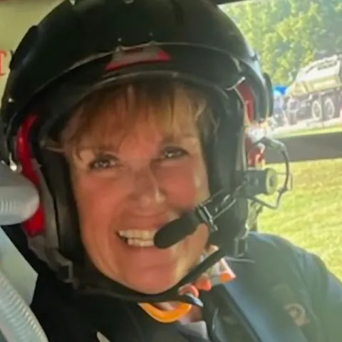 Smiling woman wearing a black aviation helmet with a built-in microphone, seated inside a helicopter with an outdoor event visible through the window in the background.