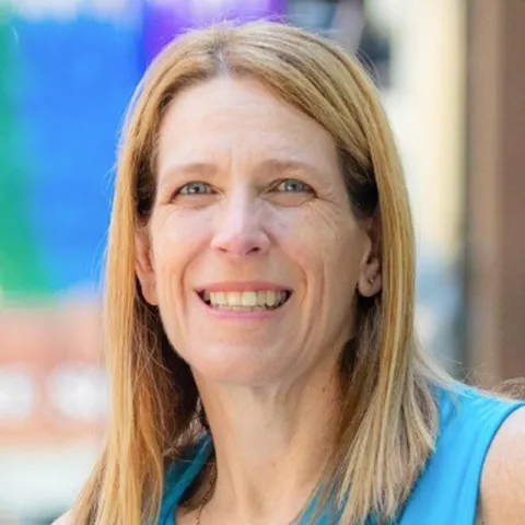 Professional headshot of a smiling woman with straight blonde hair, wearing a sleeveless blue top, set against a softly blurred outdoor background.