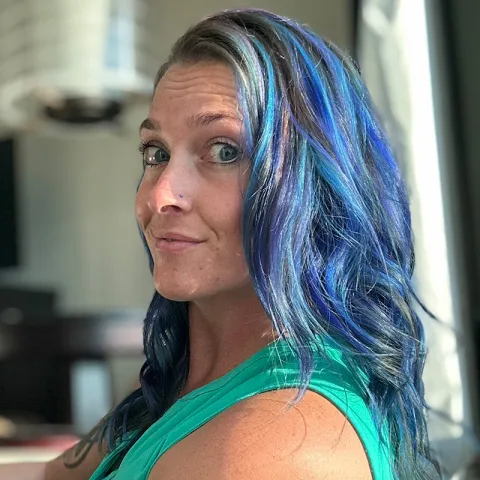 Woman with long, wavy blue and purple hair looking toward the camera with a slight smile, wearing a green sleeveless top in a well-lit indoor setting.