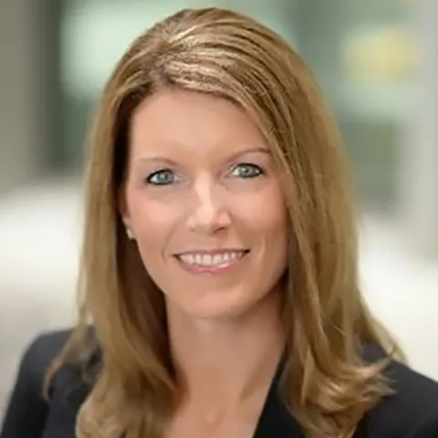 Professional headshot of a woman with long, straight, light brown hair wearing a black blazer, smiling in a softly blurred indoor background.
