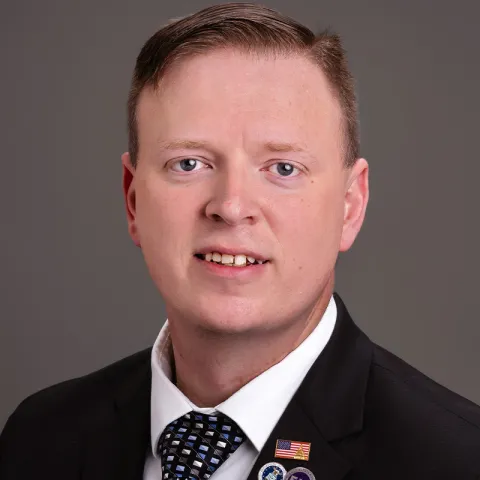 "Professional headshot of a man with short light brown hair, wearing a black suit, white shirt, and patterned tie, with multiple pins on his lapel, smiling in front of a gray background.