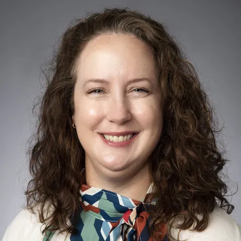 Professional headshot of a smiling woman with long curly brown hair, wearing a multicolored geometric scarf and cream sweater, against a neutral gray background.