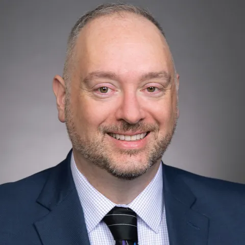 Professional headshot of a smiling man with short hair and a trimmed beard, wearing a navy suit, checkered shirt, and striped tie, against a neutral gray background.