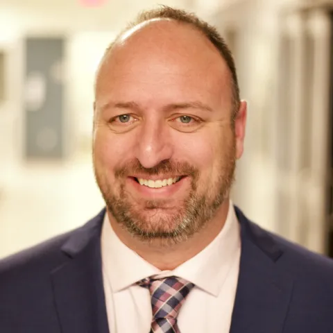 "Professional headshot of a man with a beard and short hair, wearing a navy blue suit, white shirt, and plaid tie, smiling in a brightly lit hallway.