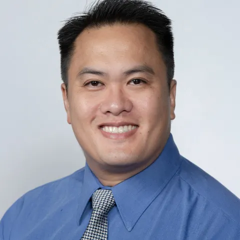 Professional headshot of a man with short black hair, wearing a blue dress shirt and patterned tie, smiling in front of a light gray background.
