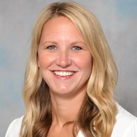 Professional headshot of a woman with long blonde wavy hair, wearing a white top, smiling in front of a light gray and blue background.