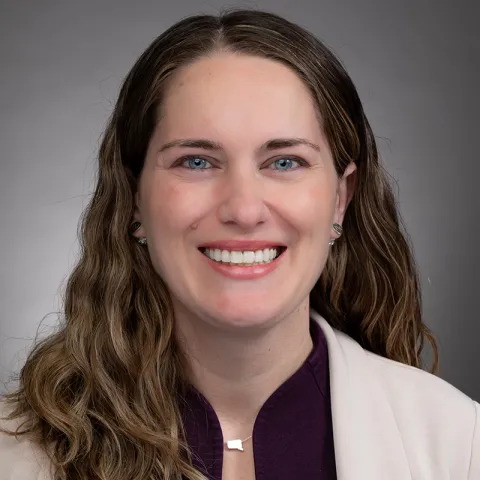 Professional headshot of a woman with long wavy brown hair, wearing a cream blazer over a dark top, smiling in front of a gray background.