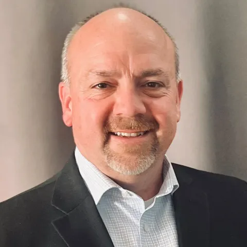Professional headshot of a smiling man with a goatee and shaved head, wearing a dark blazer over a light blue checkered shirt, posed in front of a neutral background.