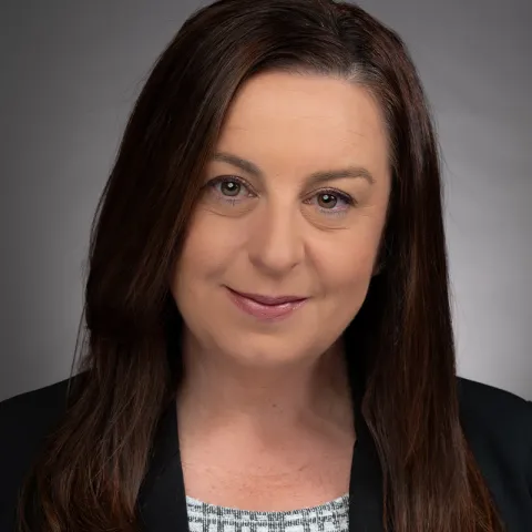 Professional headshot of a woman with long straight dark brown hair, wearing a black blazer and patterned blouse, smiling slightly against a gray background.