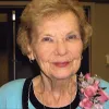 Portrait of an older woman with short light hair, wearing a light blue sweater and a floral corsage pinned on her shoulder, smiling gently at the camera.